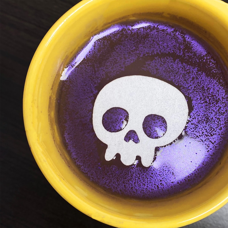 May include: A yellow ceramic mug with a purple and white skull design. The skull is white with black eyes and a black nose.