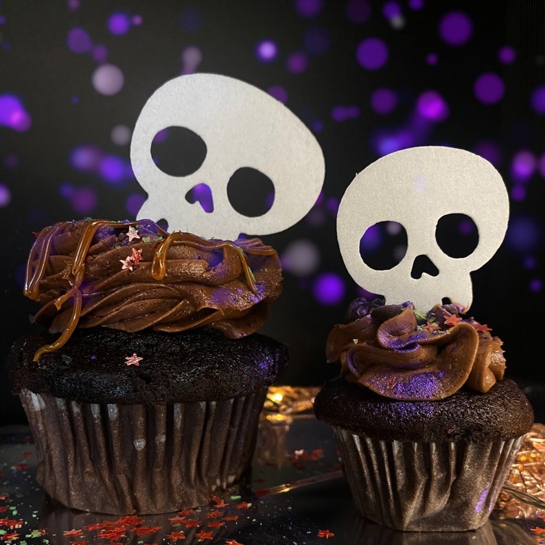 May include: Two chocolate cupcakes with brown and purple frosting decorated with sprinkles and topped with white paper skull cutouts.