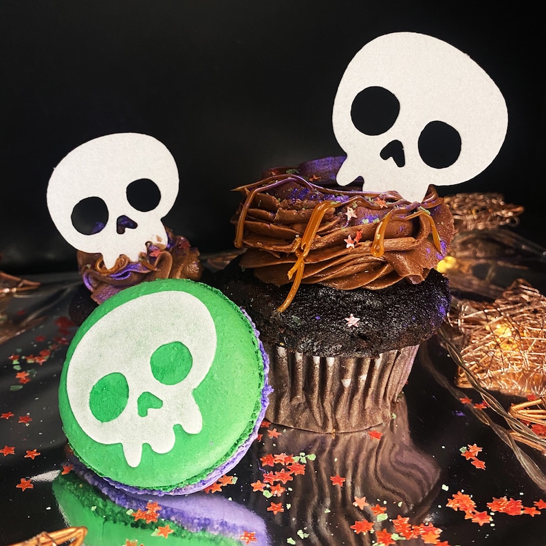 May include: Two chocolate cupcakes decorated with brown frosting and topped with white paper skull cutouts. One cupcake has a green skull-shaped macaron on top.