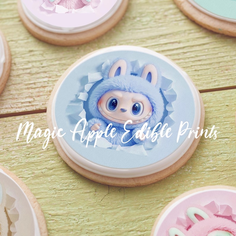 May include: Close-up of a decorated cookie with a blue cartoon character design. The character is wearing a blue hooded outfit with bunny ears. The cookie has a light blue border and the text "Magic Apple Edible Prints".