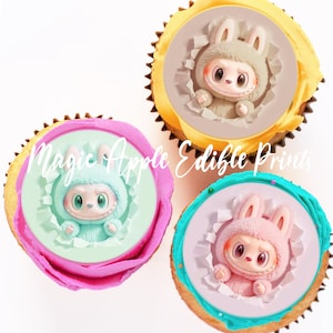May include: Three cupcakes with edible prints featuring a cartoon bunny character. The bunny is wearing a hooded outfit and peeking through a torn paper effect. The cupcakes have different colored frosting and sprinkles. The text "Magic Apple Edible Prints" is visible.