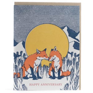 Peut inclure: Une carte de vœux illustrée de deux renards orange et blancs assis dans un champ d'épis de blé bleus. Un grand soleil jaune et des montagnes bleues se détachent sur un ciel bleu foncé. Le texte HAPPY ANNIVERSARY est écrit en rouge.
