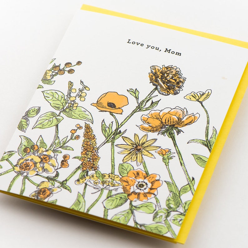 May include: A greeting card with a yellow border and a white interior featuring a floral pattern with yellow and orange flowers. The text "Love you, Mom" is printed in black at the top of the card.