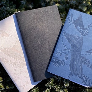 Puede incluir: Tres cuadernos con ilustraciones de pájaros en relieve. Los cuadernos son blanco, negro y azul. El cuaderno blanco tiene un pájaro blanco en una rama, el cuaderno negro tiene un pájaro negro en una rama y el cuaderno azul tiene un pájaro azul en una rama.