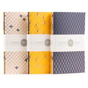 Puede incluir: Juego de tres cuadernos con cubiertas decorativas. Las cubiertas presentan diseños geométricos dorados y azul marino sobre fondos crema, amarillo y azul marino. Cada cuaderno está envuelto con una banda blanca que dice "LETTERPRESS PORCHLIGHT PRESS". Los cuadernos miden 12,7 x 20,3 cm.