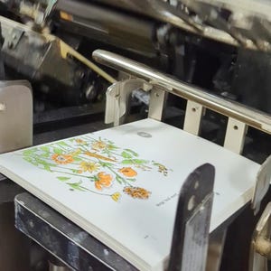 May include: A close-up of a book with a floral illustration of orange and yellow flowers on a white background. The book is being printed on a large printing press.
