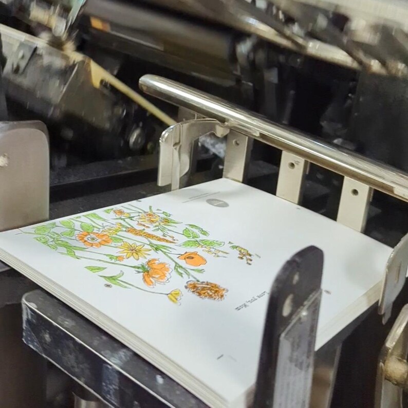 May include: A close-up of a book with a floral illustration of orange and yellow flowers on a white background. The book is being printed on a large printing press.