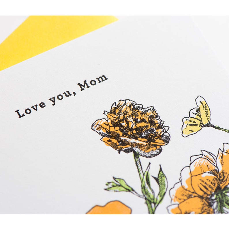 May include: A white card with the text "Love you, Mom" in black lettering. The card is decorated with a hand-drawn illustration of yellow flowers.