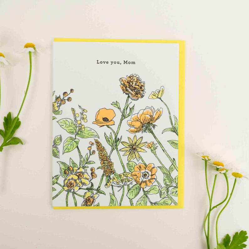 May include: A white card with a yellow border features a hand-drawn illustration of yellow and orange flowers. The text "Love you, Mom" is printed in black at the top of the card.