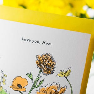 May include: A white card with a yellow border and a floral illustration. The card says "Love you, Mom" in black text.