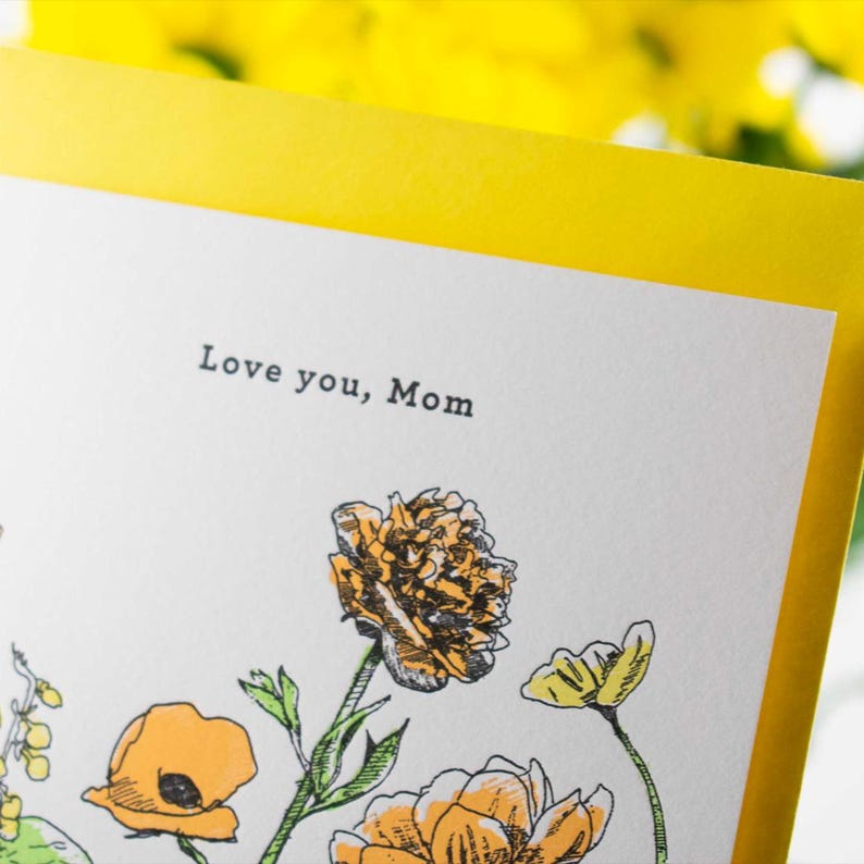 May include: A white card with a yellow border and a floral illustration. The card says "Love you, Mom" in black text.