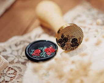 May include: A wooden handled wax stamp with a gold-colored floral design, next to a black wax seal with red roses and green leaves. The stamp and seal are on a white lace surface.