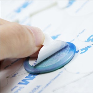May include: A hand peeling back the backing of a round, blue adhesive sticker with a white backing. The sticker is on a white surface with blue text that says "The Tape".