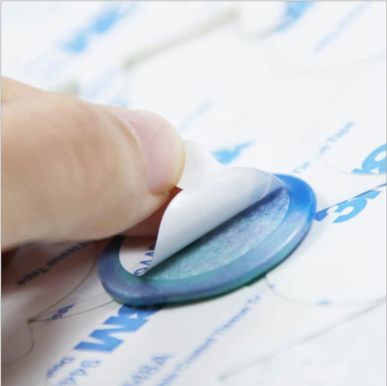 May include: A hand peeling back the backing of a round, blue adhesive sticker with a white backing. The sticker is on a white surface with blue text that says "The Tape".