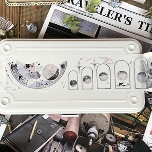 May include: A white metal box with a roll of decorative washi tape featuring a moon and star design. The tape is partially unrolled and shows a black and white illustration of a bird with wings spread.