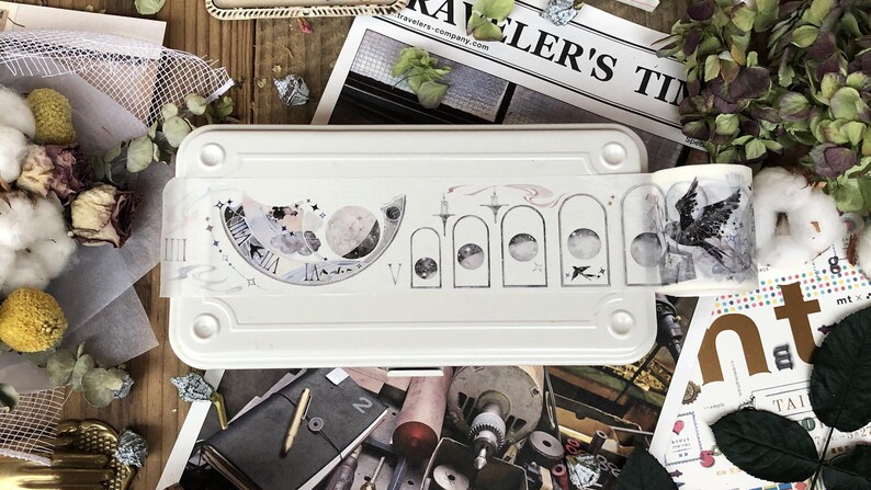 May include: A white metal box with a roll of decorative washi tape featuring a moon and star design. The tape is partially unrolled and shows a black and white illustration of a bird with wings spread.