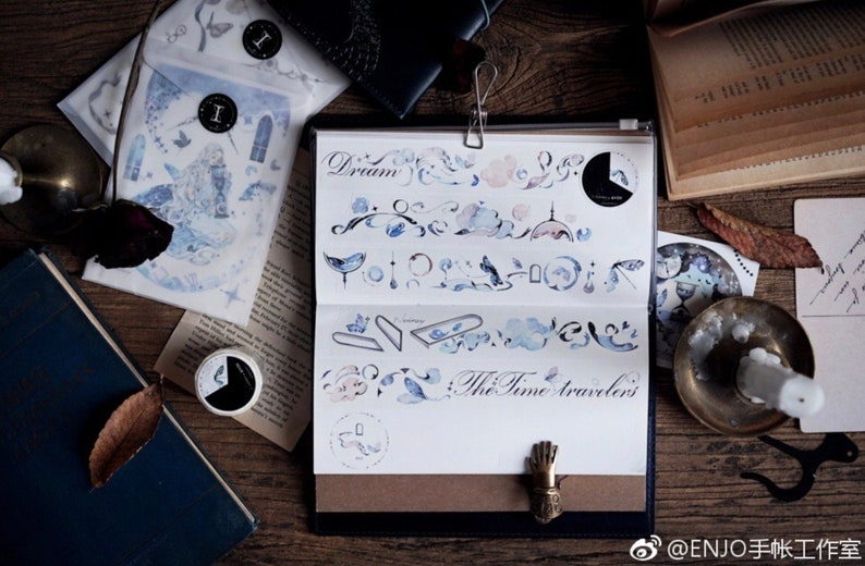 May include: A set of watercolor-style washi tape featuring a blue and white color scheme. The tape includes designs of clouds, birds, and other whimsical elements. The text "The Time Travelers" is written on the tape.