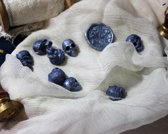 May include: A white fabric surface with a blue wax seal and several small blue skull-shaped charms.