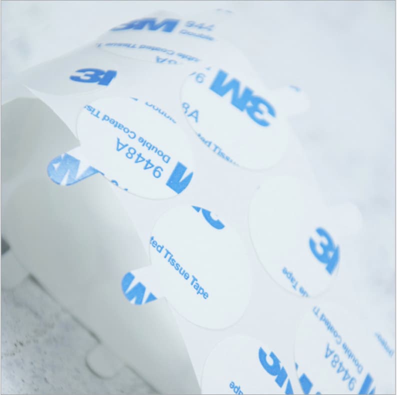 May include: A roll of white, round, adhesive stickers with blue text that reads "3M 9448A Double Coated Tissue Tape".