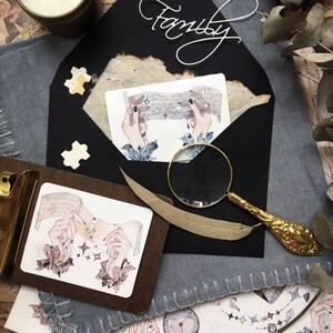 May include: A black envelope with the word "Family" written in white cursive on the front. The envelope is open and there is a watercolor illustration of hands holding a piece of paper with stars on it. There is a magnifying glass and a feather on the table next to the envelope.