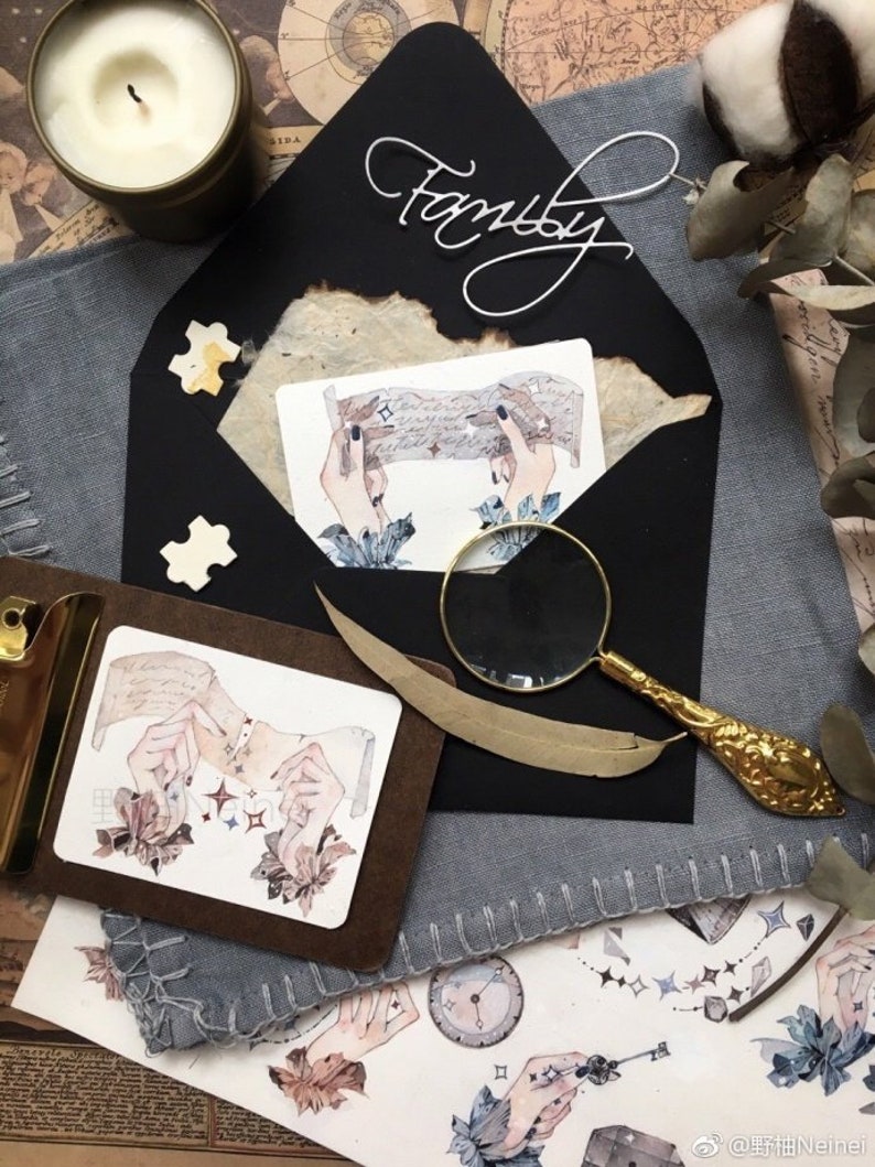 May include: A black envelope with the word "Family" written in white cursive on the front. The envelope is open and there is a watercolor illustration of hands holding a piece of paper with stars on it. There is a magnifying glass and a feather on the table next to the envelope.