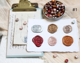 May include: Six wax seals in different colors, including silver, gold, red, pink, and copper. The seals are arranged in two rows of three on a white piece of paper. The paper is on a beige surface. There is a small tin of wax beads in the upper right corner of the image. The tin is filled with wax beads in various colors, including red, gold, and silver. The text "#1" is written in black ink on the beige surface.