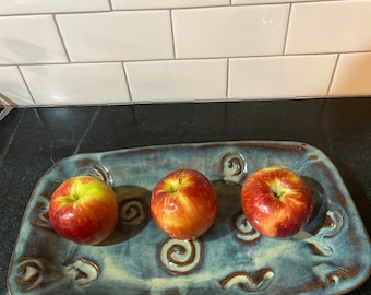 May include: Three red and yellow apples rest on a rectangular, blue-green ceramic platter. The platter features a decorative swirl design. The apples are ripe and ready to eat.