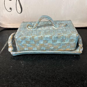 Può includere: Una burriera rettangolare in ceramica con coperchio. Il piatto e il coperchio sono azzurri con una trama intrecciata e dettagli dorati. Il coperchio ha una maniglia curva. Il piatto poggia su un vassoio abbinato.
