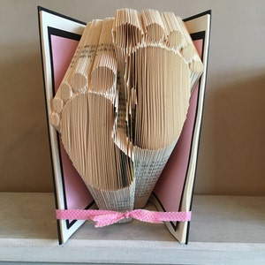 Puede incluir: Un libro doblado en forma de dos pies de bebé, con una cinta rosa atada alrededor de la parte inferior. El libro está de pie en un estante blanco.