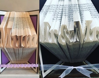 May include: Two book folding art pieces, one spelling "Mom" with a purple background and the other spelling "Love" with a black background. Both pieces are decorated with ribbon.