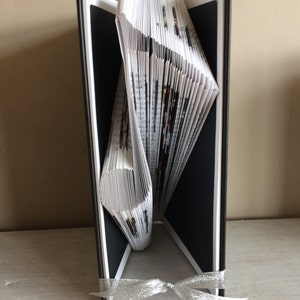 Può includere: Una scultura di arte libraria a forma del numero 7. Il libro è rilegato in nero e le pagine sono piegate per creare la forma. La scultura è appoggiata su una superficie bianca e ha un nastro argentato legato intorno.