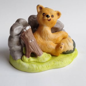 May include: A ceramic figurine of a brown bear sitting on a green grassy base with a brown tree stump and gray rocks. The bear has a happy expression and is looking to the right.
