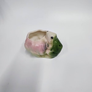 May include: A ceramic frog planter with a pink and green glaze. The frog is holding its mouth open, creating a small planter.