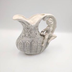 May include: A white ceramic pitcher with an ornate design and a cherub figure on the handle. The pitcher has a gray and white glaze and a detailed floral pattern.