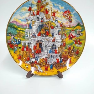 Peut inclure: Une assiette en céramique colorée représente une scène fantaisiste de chats construisant un château. Les chats portent des robes rouges et utilisent des échelles, des échafaudages et d'autres outils de construction. Un chat en ballon à air chaud survole la scène. L'assiette a un bord doré et est exposée sur un support en bois.