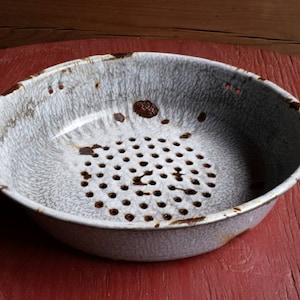 Puede incluir: Un colador de esmalte blanco con manchas de óxido marrón y un patrón de agujeros en el fondo. El colador está sobre una superficie de madera roja.