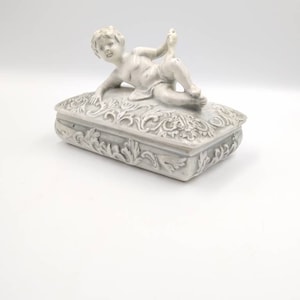 May include: A white ceramic trinket box with a cherub figurine on the lid. The box is decorated with a floral design.