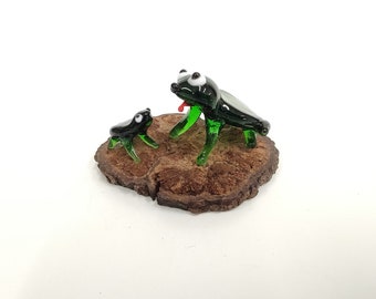 May include: Two glass frog figurines on a wooden base. The larger frog is green with white eyes and a red tongue. The smaller frog is black with white eyes.