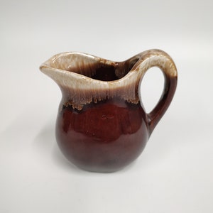 Pode incluir: Um pequeno creamer de cerâmica marrom e branco com um corpo arredondado e uma alça. O creamer tem um acabamento esmaltado com uma base marrom e uma borda branca.