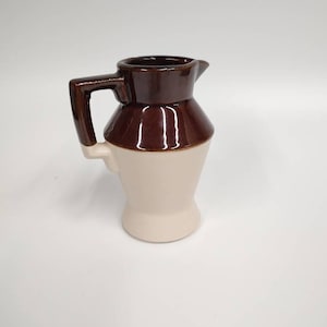 Puede incluir: Una pequeña jarra de cerámica marrón y blanca con asa. La jarra tiene un esmalte marrón en la mitad superior y un esmalte blanco en la mitad inferior.