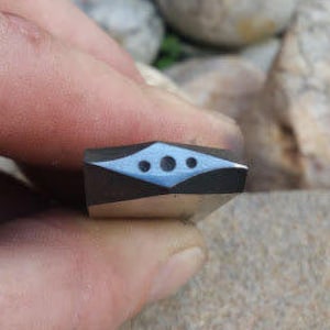 Puede incluir: Pequeño objeto rectangular con base negra y diseño geométrico azul claro y blanco. El diseño incluye una forma de diamante central con tres pequeños círculos. El objeto está en la mano de una persona.