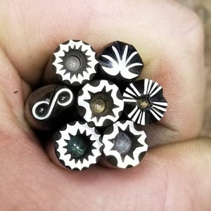 May include: A collection of metal stamps with various geometric and floral designs. The stamps have black and white patterns, including starbursts, sunbursts, and abstract shapes. The stamps are held in a hand, showcasing the detailed designs.