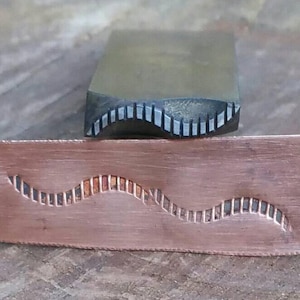 Puede incluir: Una herramienta para trabajar metales y una placa de cobre. La herramienta tiene un borde estampado y la placa de cobre tiene un diseño ondulado a juego. Las herramientas se utilizan para estampar metales y crear patrones decorativos.