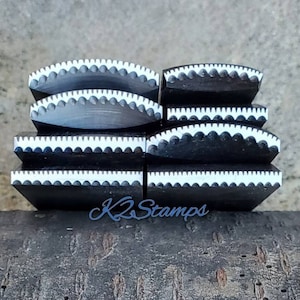 Può includere: Una collezione di timbri in legno neri con bordi smerlati bianchi. I timbri sono impilati su una superficie di legno, con il testo blu "K2 Stamps" visibile. Questi timbri sono probabilmente utilizzati per progetti di artigianato.