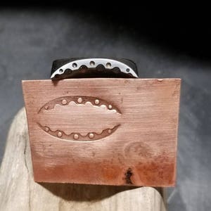 以下が含まれることがあります： 目のようなデザインの銅製金属スタンプと、上部に銀色の金属片。スタンプは長方形で、表面はざらざらしており、木片の上に置かれています。
