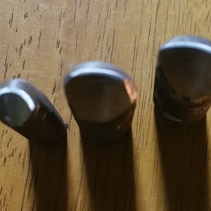 May include: Three metal golf club heads with a white stripe on the top edge, arranged in a row. The clubs are dark in color and appear to be made of metal. The background is a wooden surface.