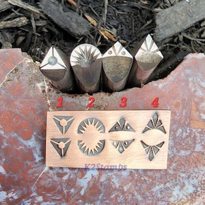 May include: Four metal stamps with geometric designs, including triangles, sunbursts, and diamond shapes. A wooden block displays stamped impressions of the same designs. The text "K2Stamps" is visible on the block.