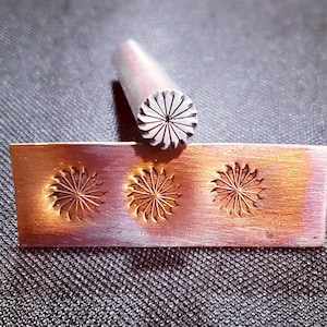May include: A metal stamping tool with a floral design, next to a copper plate with three stamped impressions of the same pattern. The tool is cylindrical with a detailed, radial design. The copper plate shows the stamped impressions.