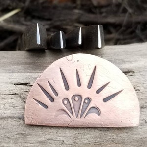 Peut inclure: Un pendentif en demi-cercle de couleur cuivrée avec un motif de soleil et trois petits objets rectangulaires foncés avec des accents blancs. Le pendentif est posé sur une surface en bois vieilli.