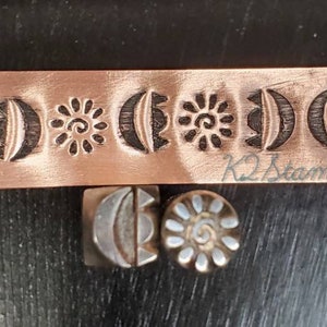 May include: Copper metal stamps with celestial designs, including moons and suns. A long copper strip features repeating patterns. Two individual stamps, one square and one round, are also visible. The text "K2Stamps" is on the copper strip.